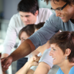 Male teaching a woman by pointing to something on her screen, both are laughing. There are others in the background.